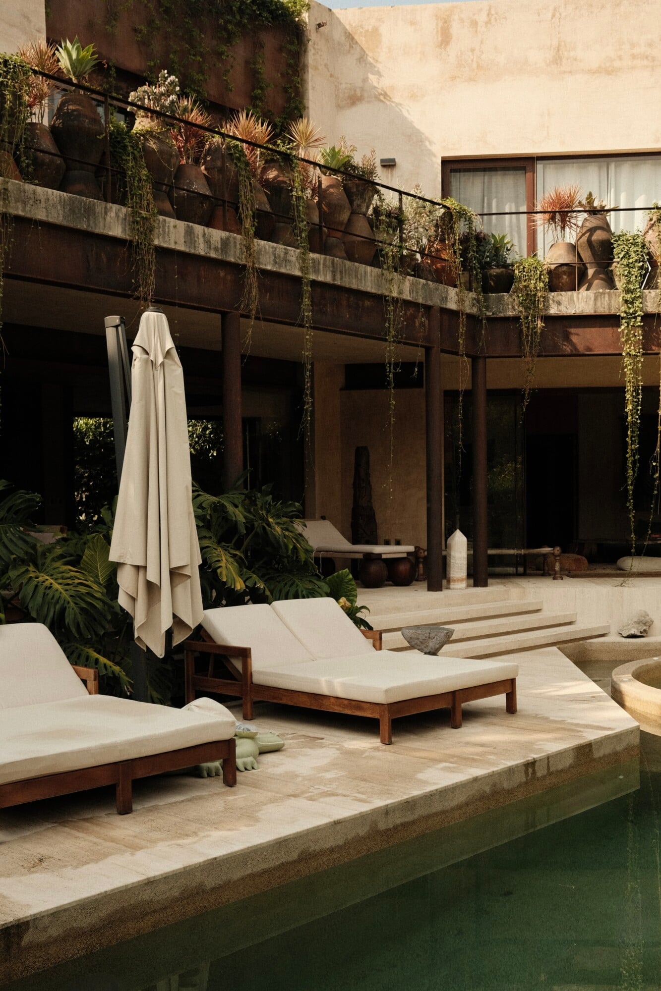 Casa Lumeria, Mexico — Courtyard with limestone pool, wooden loungers, cream umbrellas, cascading vines on brutalist concrete architecture, minimalist luxury design.